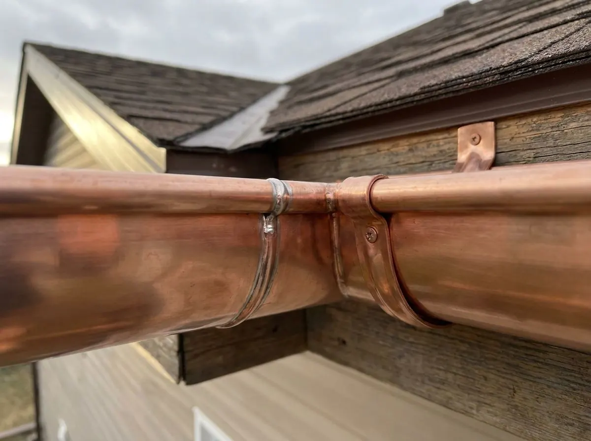 Close-up of a half-round copper gutter showing a neat soldered seam between two sections and a copper bracket supporting the gutter beneath.