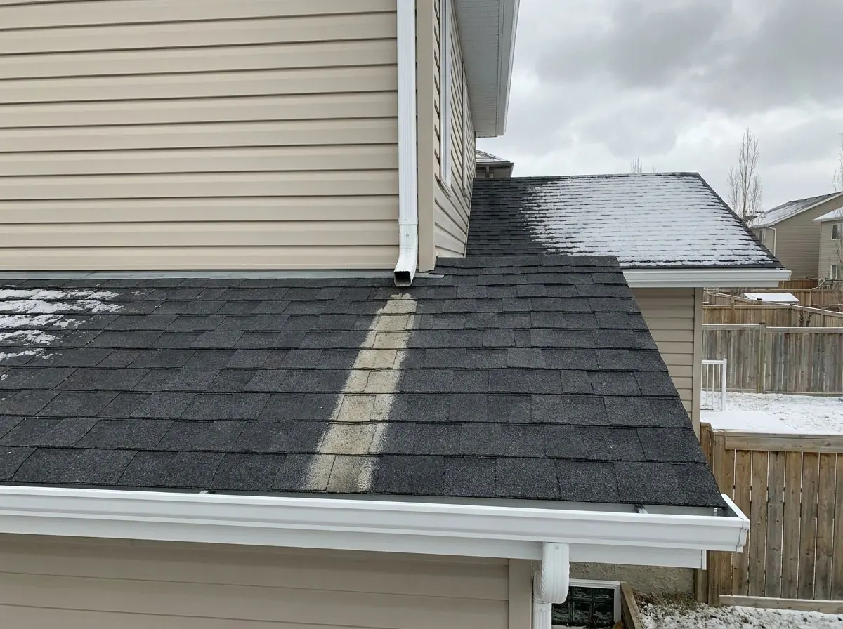 Close-up of a lower roof where water from an upper downspout has washed a pale stripe of granules off the dark shingles down toward the eavestrough.
